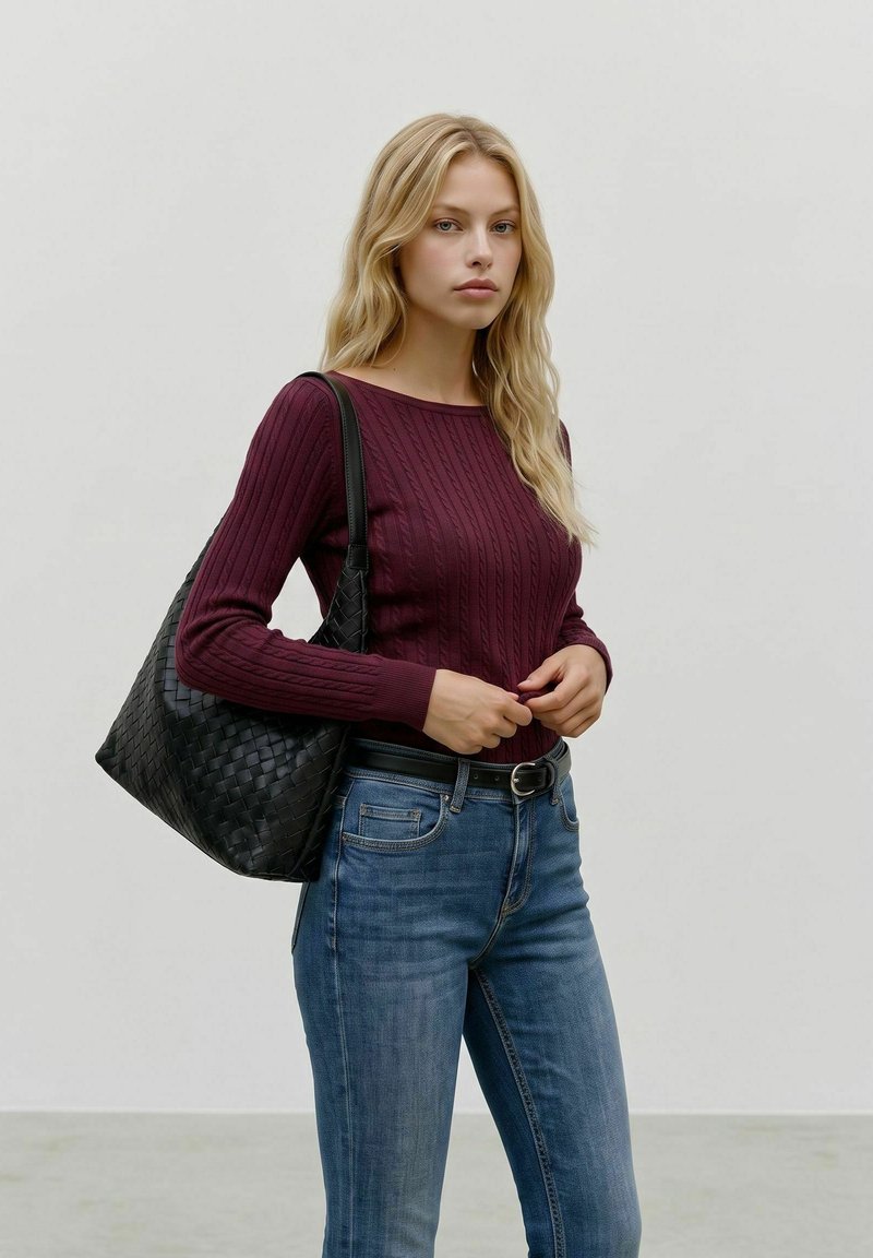 Jeune femme aux cheveux blonds porte un pull bordeaux et un jean bleu, portant un grand sac à bandoulière en cuir tressé noir.