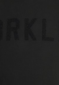 T-shirt in cotone nero con letteratura testurizzata e in rilievo che scrive "BRKLYN" in un colore nero abbinato. Superficie liscia con una finitura morbida.