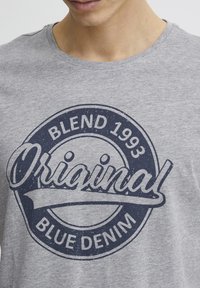 Pilkas medvilnės marškinėliai su tamsiai mėlynu apvaliu logotipu, kuriame užrašyta "Blend 1993" ir "Original Blue Denim", matoma tekstūra ir sutrūkinėjusi detalizacija.