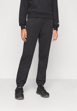 Person iført sorte sweatpants og matchende sort sweatshirt, stående på en ensfarvet hvid gulv og baggrund.