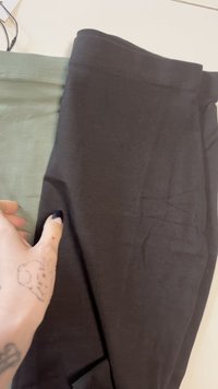 Mână cu tatuaj de iepure ținând o țesătură pliată maro închis și verde măsliniu pe o suprafață albă.