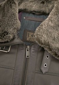 Braune Lederjacke mit einem Kunstpelzkragen, sichtbarem Reißverschluss und einem Denim-Futter mit einer Logoplatte. Detaillierte Nähte und metallische Akzente.