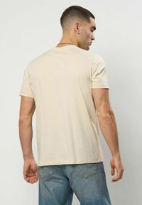 Beige kortermet t-skjorte laget av mykt stoff. Har rund hals og rett snitt, med jevn tekstur og minimal detaljering.