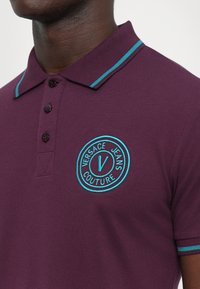 Κοντινό πλάνο ενός σκούρου δέρματος ατόμου που φοράει ένα μωβ πόλο μπλουζάκι με τουρκουάζ ρίγες στο γιακά και στα μανίκια, το οποίο φέρει ένα κυκλικό λογότυπο της Versace Jeans Couture.