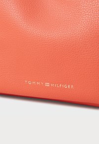 Bolso de mano de cuero coral con superficie texturizada, que presenta el logo dorado "TOMMY HILFIGER". Forma compacta, diseño minimalista.