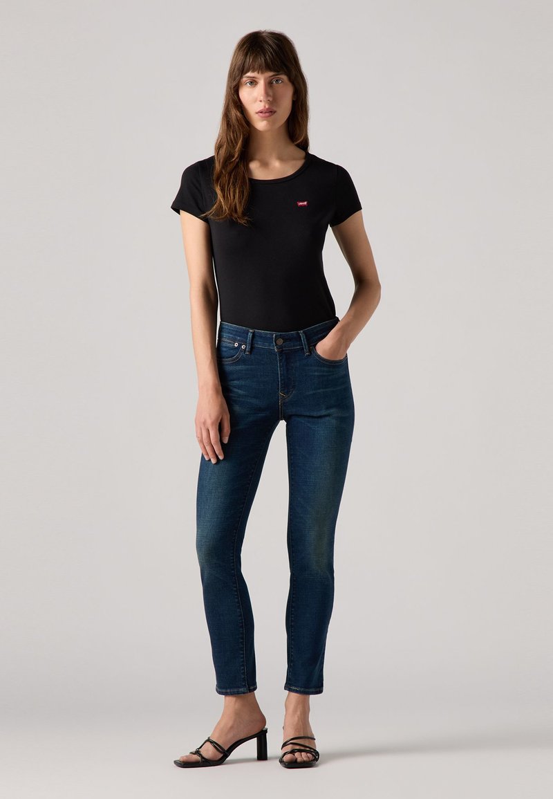 Schwarzes Baumwoll-T-Shirt mit kurzen Ärmeln und rotem Logo, kombiniert mit dunklen blauen Slim-Fit-Jeans. Das Model trägt schwarze Sandalen mit Riemchen und steht vor einem neutralen Hintergrund.
