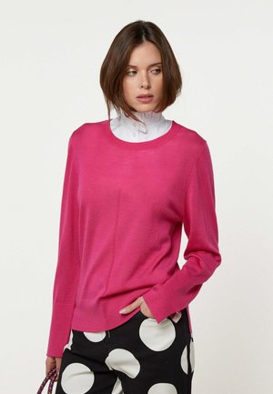 Vrouw draagt een felroze trui over een wit met knopen sluitend overhemd, zwarte broek met grote witte stippen, houdt een handtas met kralen vast.