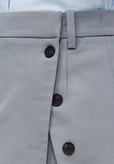 Κοντινό πλάνο γκρι παντελονιού με τρία μαύρα κουμπιά μερικώς κουμπωμένα και μια θηλιά για ζώνη πάνω από τη μέση, φορεμένο πάνω από πουκάμισο με μπλε ρίγες.