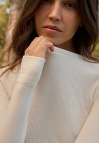 Világos bézs, ribbed anyagú, hosszú ujjú felső, magas nyakkal. Közeli felvétel az anyag textúrájáról és a varrás részleteiről. Puha és testhezálló kialakítás.