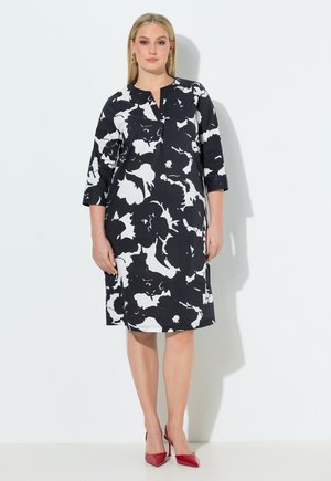 Kvinde iført sort og hvid knælængde kjole med blomsterprint og trekvartærmer samt røde højhælede sko, stående mod en hvid baggrund.