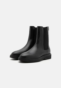 Botines de cuero negro que cuentan con una textura suave, puntera redondeada, paneles laterales elásticos y una suela negra baja y gruesa.