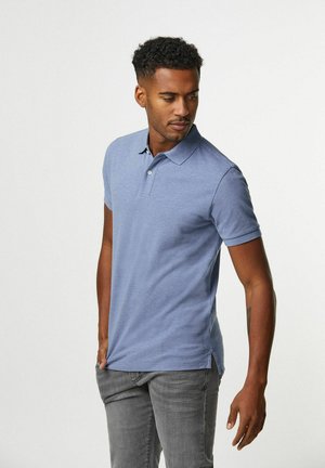 Hellblaues Poloshirt mit klassischem Kragen, kurzen Ärmeln, seitlichen Schlitzen und dezentem Strukturmuster. Kombiniert mit grauen Jeans.