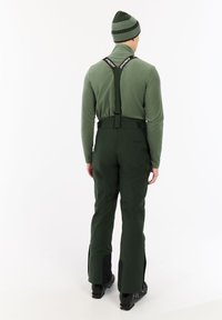 Man in een groene beanie, een groen langemouwtop, donkergroene skibroek met bretels, en zwarte skischoenen, staande met de rug naar de camera op een witte achtergrond.