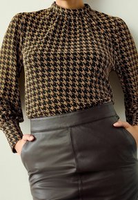 Haut à manches longues en tissu noir avec un motif pied-de-poule doré, col montant et détails froncés, associé à une jupe en cuir noire.