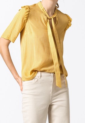 Blusa gialla a fantasia con maniche corte a volant e scollatura annodata, realizzata in tessuto morbido, abbinata a pantaloni di colore chiaro.