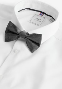 Fehér, texturált felületű ing, sötétszürke masnival. A gallér friss, és a címke a "NEXT TAILORING" feliratot viseli.