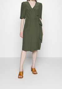 Olive green wrap dress with short puff sleeves, two front pockets, and a tie detail at the waist. Paired with brown high-heeled sandals.