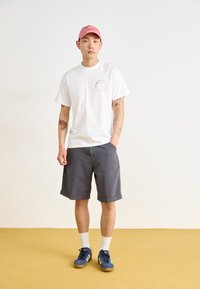 White cotton t-shirt with a small red graphic, gray shorts, blue sneakers with white accents, and a pink cap. Plain background.