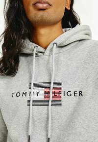 Osoba w jasnoszarej bluzie z kapturem, mająca na piersi czarno-czerwony pasek z logo Tommy Hilfiger, pokazująca dolną część twarzy i długie ciemne włosy.