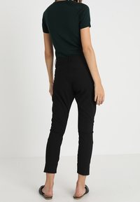 Femme portant un pantalon noir ajusté à la cheville, un haut vert foncé à manches courtes et des chaussures noires à enfiler, se tenant contre un fond blanc.