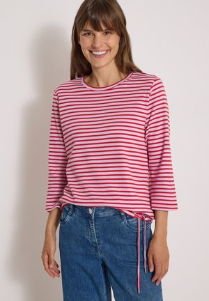 Femme souriante portant un haut à manches longues rayé rouge et blanc avec un jean bleu, debout devant un fond clair uni.