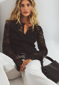 Black lace cardigan with long sleeves and button closure, worn over a dark top, paired with light-colored pants and a black shoulder bag.