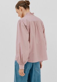 Donna con i capelli raccolti in uno chignon, che indossa una blusa rosa larga con maniche arricciate e jeans di denim blu, rivolta di spalle su uno sfondo neutro.