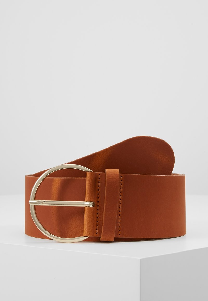 Ceinture en cuir marron avec une grande boucle ronde en or, présentée enroulée sur une surface blanche sur un fond gris uni.