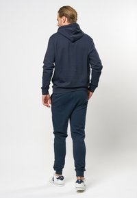 Mørkeblå hettegenser med snøring i hetta, i kombinasjon med matchende joggebukser. Materialet ser mykt ut med ribbede mansjetter. Skoene inkluderer hvite joggesko.