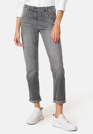 Kvinde iført op rullede grå jeans med forlommer og hvide sneakers, der står på en ensfarvet baggrund.