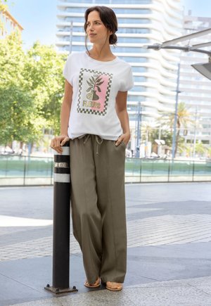 White t-shirt featuring a floral graphic with green and pink accents, paired with loose olive trousers and tan sandals, standing by a pole.
