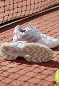 Witte tennisschoenen op een gravelbaan vlak bij een net, eentje toont een patroon op de zool, met een gedeeltelijk zichtbare gele tennisbal.