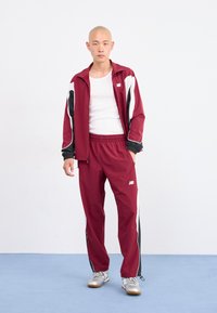 Bordeaux tracksuit med hvide og sorte detaljer, som består af en zip-jakke og bukser med elastisk talje. Sølvfarvede sportssko fuldender looket.