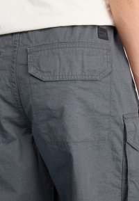 Grå cargo-byxor med en strukturerad textil, med sidofickor och en ficka bak, markerad med en svart etikett.