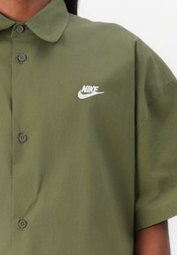 Olive green short-sleeve button-up shirt made of smooth fabric, featuring a white Nike logo on the upper left side and two buttons visible.