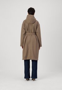 Manteau beige ceinturé avec capuche, doté d'une coupe décontractée et de manches longues, associé à un jean large bleu foncé et des talons pointus.