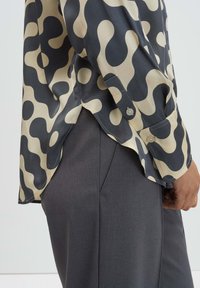 Flowy button-up shirt in dark gray with beige abstract shapes, featuring a curved hem and long sleeves. Paired with gray tailored trousers.