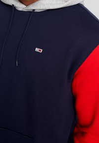 Sudadera con capucha que presenta un cuerpo azul marino, mangas rojas y una capucha gris. Incluye un pequeño logo en el pecho y detalles con cordón.