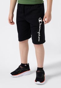Enfant portant un short noir Champion et des baskets noires Champion avec des accents orange et blancs, se tenant sur un fond blanc.