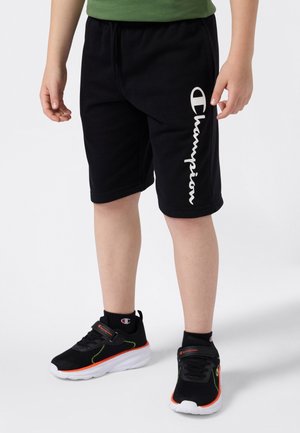Kind, der schwarze Champion-Shorts und schwarze Champion-Sneaker mit orangefarbenen und weißen Akzenten trägt, steht vor einem weißen Hintergrund.