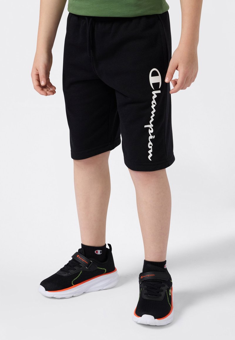 Enfant portant un short noir Champion et des baskets noires Champion avec des accents orange et blancs, se tenant sur un fond blanc.