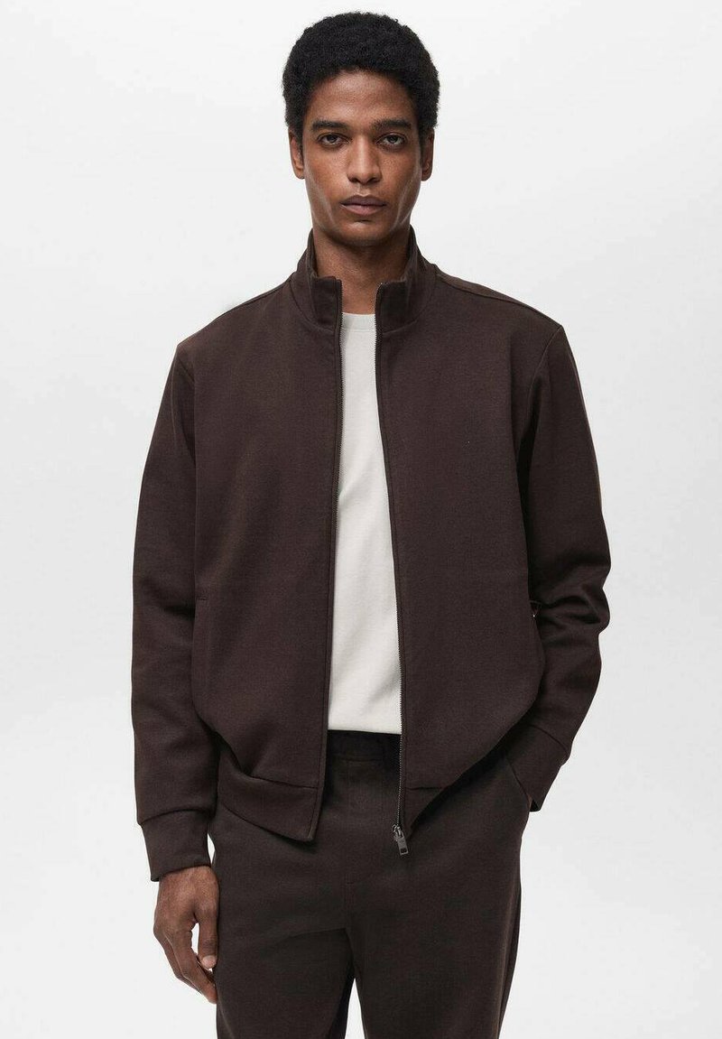 Giacca marrone con zip e colletto alto, realizzata in materiale texture. Indossata sopra una camicia chiara. Presenta tasche laterali e una vestibilità rilassata.