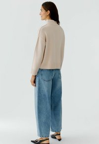 Hellbeige Pullover mit hohem Rippkragen, kombiniert mit lockeren blauen Jeans und schwarzen Schuhen. Stricktextur, lässiger Schnitt.