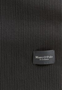 Zwarte ribbelgebreide stof met een rechthoekig label erop gestikt, met de tekst "Marc O'Polo EST. IN STOCKHOLM" in witte letters.