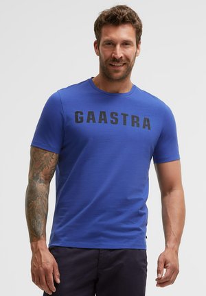 Man with short brown hair and a beard wears a blue Gaastra t-shirt and dark pants, showing a tattooed right arm.