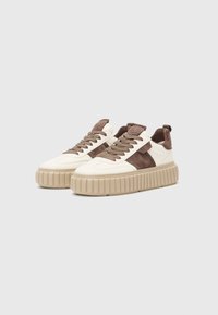 des baskets avec une tige en cuir crème, des accents en daim marron, une semelle en caoutchouc beige épaisse avec une texture nervurée, et des lacets marron.