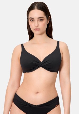 Mujer con un traje de baño de dos piezas negro con detalle de tela torcida en la parte superior e inferior, de pie contra un fondo claro y liso.