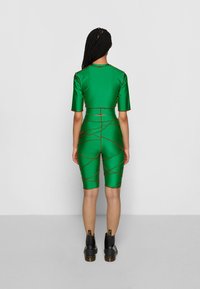 Conjunto verde ajustado com mangas curtas e calções de ciclista, apresentando costura contrastante em vermelho num padrão cruzado. Usado com botas pretas.