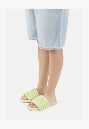 Sandales en caoutchouc vertes avec un design supérieur nervuré. Semelle plate visible et bout ouvert. Portées avec des shorts en denim clairs.