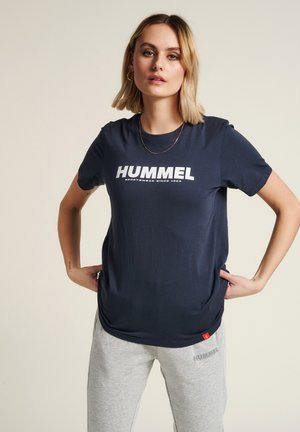Marineblå t-shirt med hvid "HUMMEL" logo. Korte ærmer, rund halsudskæring, blødt stof. Grå sweatpants med "HUMMEL" tekstlogo på venstre lår.